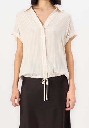 Kobieta ubrana w beżową, jasną, paskowaną bluzkę na guziki z krótkim rękawem i sznurkiem u dołu, w połączeniu z czarną spódnicą.