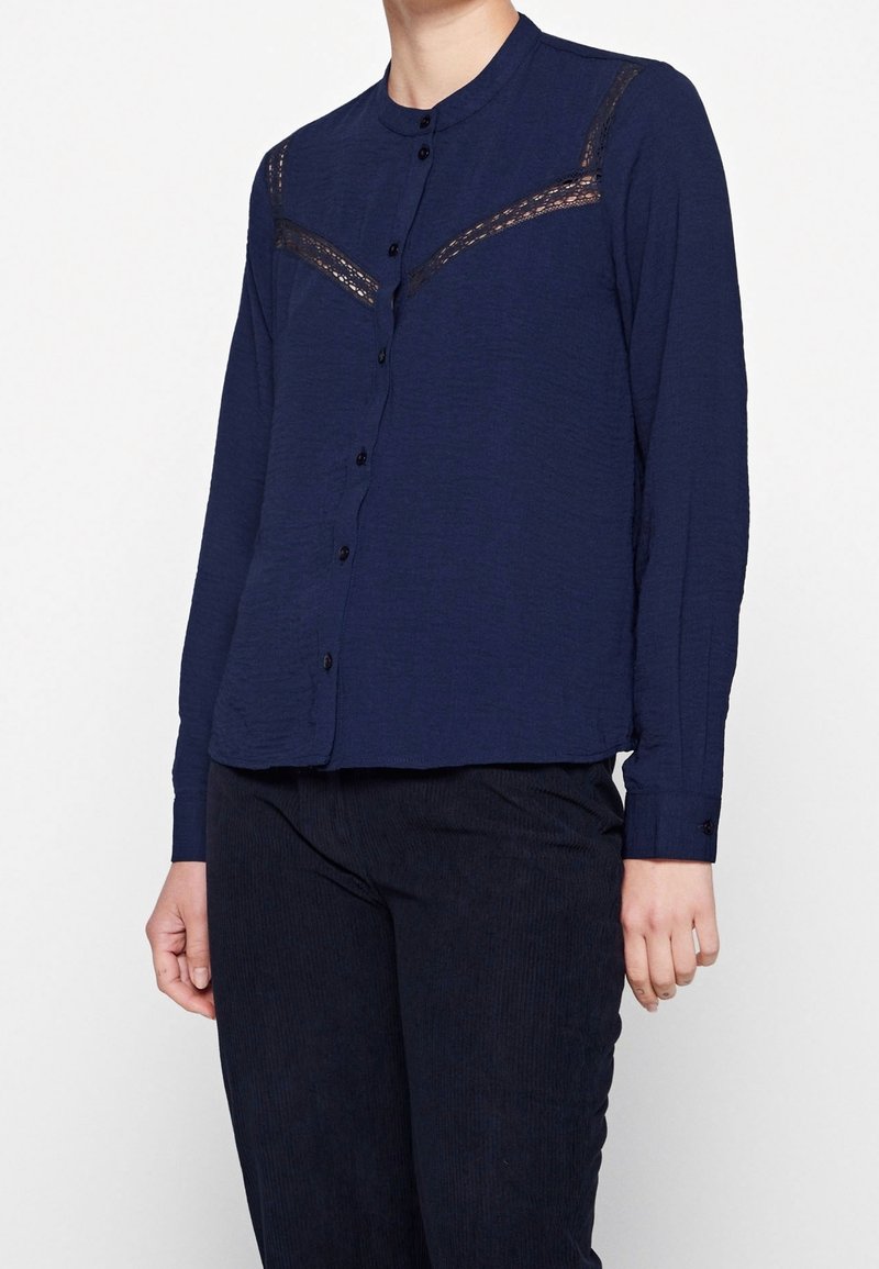 Femme portant une chemise bleu marine à manches longues boutonnée avec des détails en dentelle sur la poitrine, associée à un pantalon sombre.