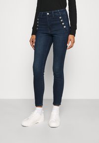 Mörkblå högmidjade skinny jeans med silverknappdetaljer på sidorna, tillverkade av denim, i kombination med vita sneakers och strumpor.