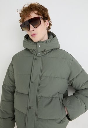 Grüne Pufferjacke mit hohem Kragen, Druckknöpfen und Seitentaschen. Das Model trägt große, dunkle, rechteckige Sonnenbrillen. Glatte Textur.