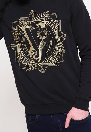 Sweat-shirt noir avec grand logo doré Versace Jeans affichant un "VJ" stylisé et un motif complexe de lion dans un cercle à motif géométrique.