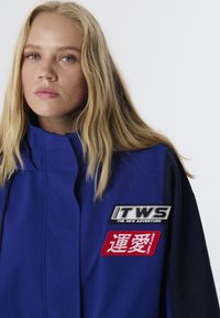 Blaue Jacke mit hohem Kragen, schwarzen Ärmeln und zwei bestickten Aufnähern: einer in Schwarz-Weiß, der andere rot mit weißem Text.