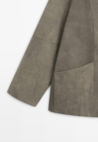 Manica di giacca in suede grigio e sezione inferiore del torso con una grande tasca a toppa visibile e dettagli delle cuciture su sfondo bianco.