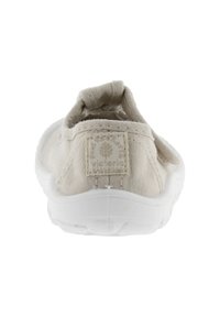 Zapato slip-on de lienzo beige con suela de goma blanca, que cuenta con un parche de logo tejido y un respaldo texturizado. Diseño ligero y flexible.