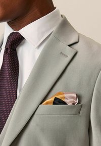 Chaqueta de traje gris claro con solapa muesca, camisa de vestir blanca y una corbata texturada color marrón burdeos. El pañuelo de bolsillo presenta patrones geométricos en amarillo, negro y rosa.