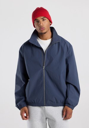 Jonge man met een rode beanie, marineblauwe zip-up jas, wit shirt en lichtgrijze joggingbroek, staand tegen een effen achtergrond.