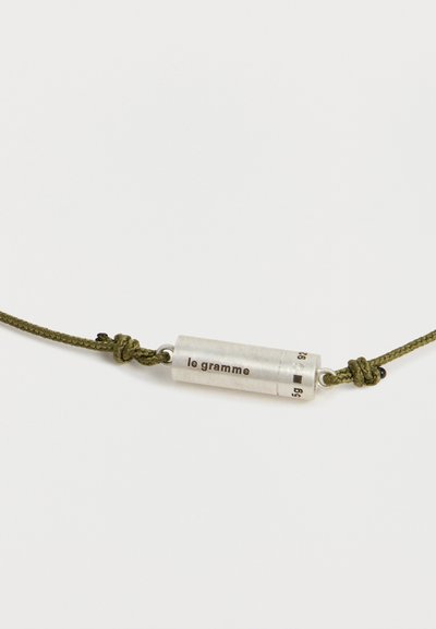 Pendentif cylindrique en argent gravé de "le gramme" attaché à un cordon vert olive avec des nœuds de chaque côté.