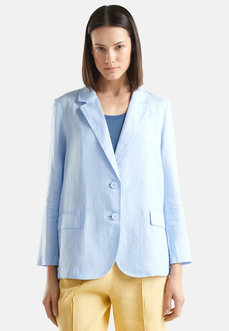 Donna che indossa un blazer azzurro chiaro con bottoni sopra una maglia blu e pantaloni gialli larghi, in piedi su uno sfondo bianco semplice.