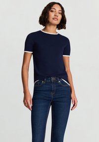 Marineblaues T-Shirt mit kurzen Ärmeln und weißem Besatz am Halsausschnitt und an den Ärmeln, kombiniert mit dunkelblauen Jeans. Glatter Stoff, figurbetontes Design.
