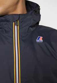 Marinefarbene Windbreaker-Jacke mit hohem Kragen, einem bunten vertikalen Reißverschluss mit roten, gelben und weißen Streifen sowie einem K-Way-Logo-Patch.