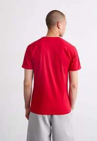 Červené bavlněné tričko s krátkými rukávy a kulatým výstřihem, hladká textura, přiléhavý design, zobrazeno zezadu s šedými tepláky.