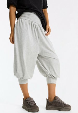 Personne portant un pantalon ample court gris clair et des baskets gris foncé, debout devant un fond blanc uni.