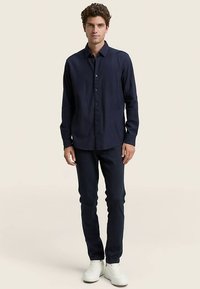 Camicia a maniche lunghe blu navy con chiusura a bottoni, design aderente, colletto, abbinata a pantaloni scuri e sneaker bianche su sfondo chiaro.