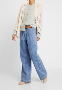 Blazer beige clair porté sur un cardigan vert menthe à boutons, associé à un pantalon en denim bleu ample avec une ceinture marron clair, le tout complété par des chaussures nude.