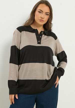 Kvinde iført en løs, langærmet polo med brede, horisontale sorte og beige striber, kombineret med blå jeans, poserer foran en ensfarvet baggrund.