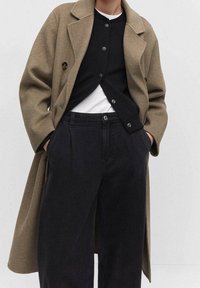 Cappotto lungo verde oliva in misto lana con colletto a punta, abbinato a un cardigan nero con bottoni, e pantaloni scuri larghi.