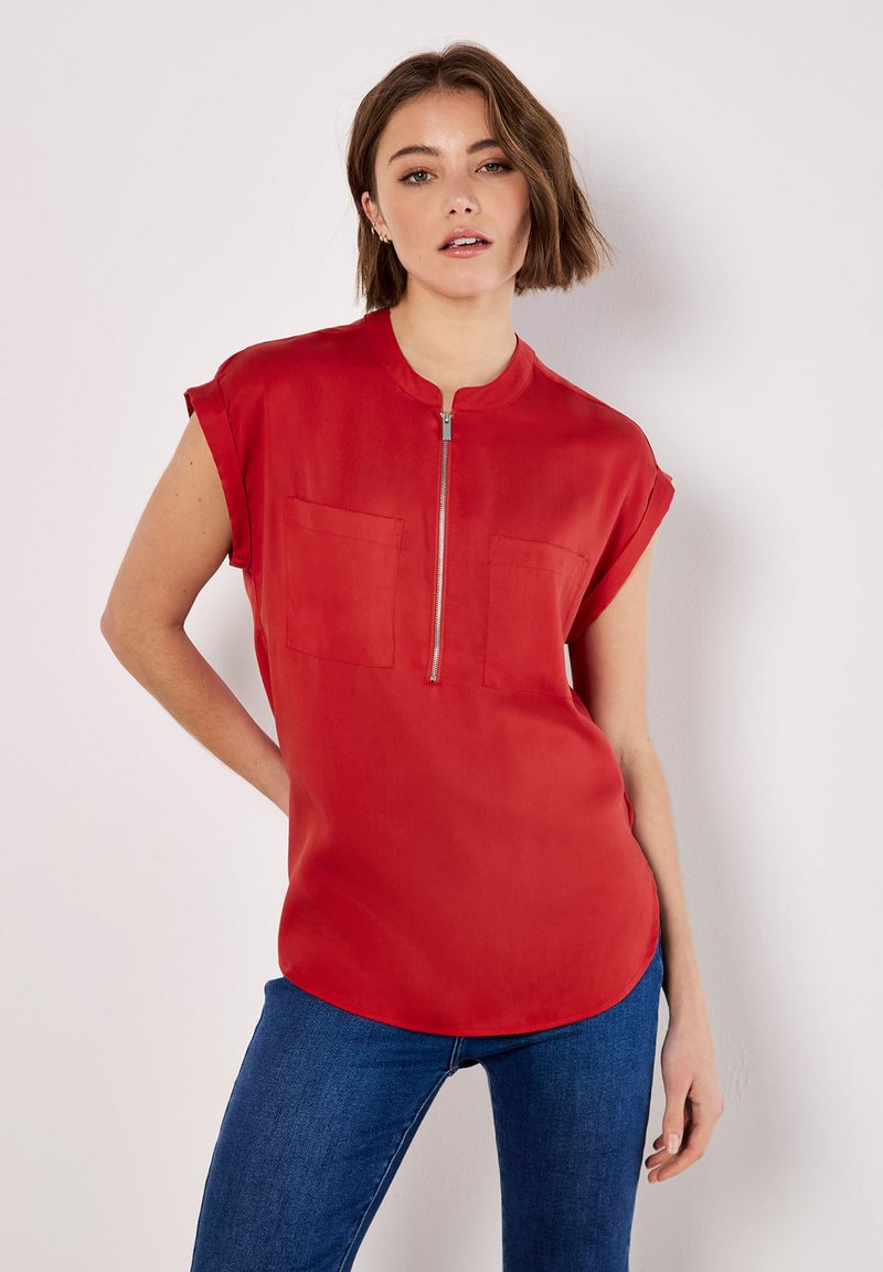 Rote Bluse mit kurzen Ärmeln und hohem Kragen, ausgestattet mit einem Frontreißverschluss und zwei Brusttaschen; glattes Material, lockere Passform und abgerundeter Saum.