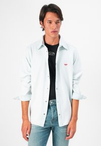 Joven vestido con una camisa de mezclilla azul clara sobre una camiseta negra y jeans azules, de pie frente a un fondo blanco.