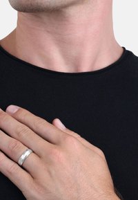 Un hombre con una camisa negra se coloca la mano sobre el pecho, llevando un anillo de boda de plata en la mano izquierda, expresando un gesto solemne.