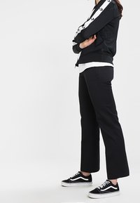 Black zip-up athletic jacket with white stripes and logos on sleeves, paired with black wide-leg pants and black sneakers.