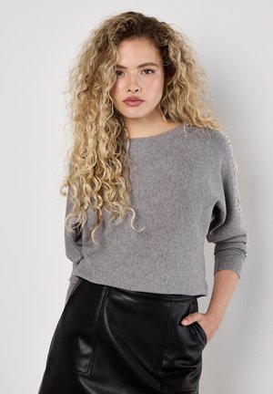 Grauer Strickpullover mit Rundhalsausschnitt und Dreiviertelärmeln, verziert mit Perlen entlang der Ärmelnähte. Kombiniert mit einem schwarzen Lederrock.