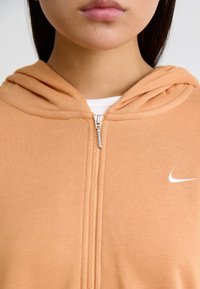 Zip-up hoodie i persikofärg, tillverkad av mjukt material, med en vit Nike-logotyp på bröstet och en dragsko i huvan.