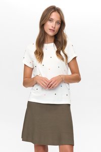 Junge Frau in einem weißen T-Shirt mit Punkten und einem olivgrünen Rock, die mit locker gefalteten Händen vor einem weißen Hintergrund steht.