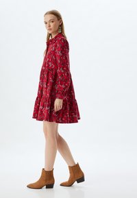Rotes Paisley-Musterkleid mit langen Ärmeln und gestuftem Rock. Kombiniert mit braunen Ankle Boots mit gerippten Details und Blockabsatz.