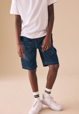 Person iført hvide sneakers, hvide sokker med sorte striber, jeansshorts og en hvid t-shirt med korte ærmer mod en beige baggrund.