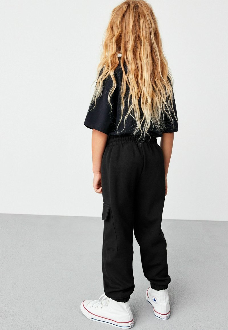 Enfant aux longs cheveux blonds portant un t-shirt noir, un pantalon cargo noir et des baskets blanches montantes, de dos sur un sol gris.