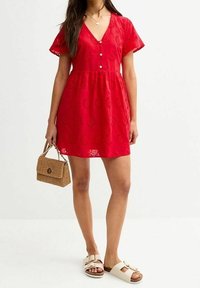 Czerwona sukienka z krótkimi rękawami, dekoltem w kształcie litery V, z detalami w postaci guzików i haftem kwiatowym. Noszona z beżowymi sandałami na płaskiej podeszwie oraz trzymana w plecionej torebce.