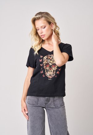 Blonde vrouw met golvend haar kijkt omlaag, draagt een zwart T-shirt met een bloemige schedel en vlindersontwerp en grijze spijkerbroeken.