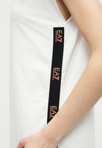 Άσπρο μανίκι τοπ χωρίς μανίκια με κάθετη μαύρη λωρίδα που διαθέτει επαναλαμβανόμενο πορτοκαλί λογότυπο "EA7 Emporio Armani" κατά μήκος της πλαϊνής ραφής.