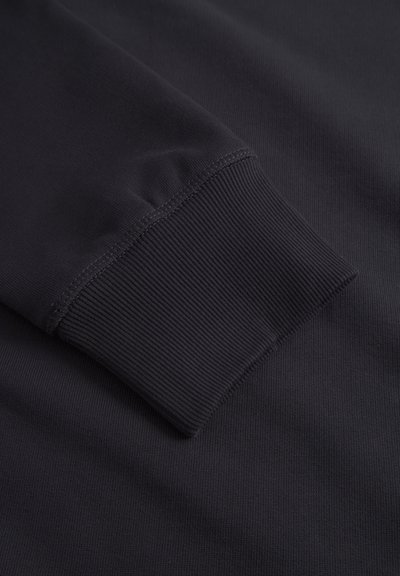 Povečan posnetek rokava črne jopice, ki prikazuje šive rebrastega manšeta in teksturo tkanine.