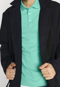 En person som bär en svart kappa över en mintgrön polo skjorta, och håller kappa kanterna med båda händerna i midjehöjd.