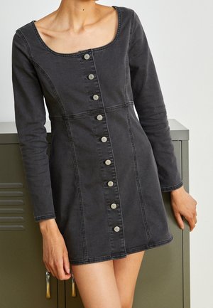 Femme portant une robe en denim noire ajustée à manches longues avec fermeture à boutons sur le devant, debout à côté d'un casier vert.