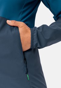 Veste bleue et bleu foncé avec une poche zippée ; texture lisse ; poignets élastiques ; présente une étiquette verte pour le branding.