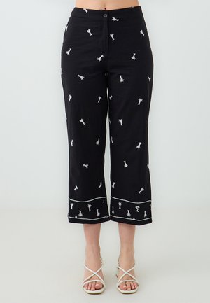 Schwarze, kurze Hosen mit weißen bestickten Palmbaum-Motiven und einem kontrastierenden weißen Saumabschluss. Das Material wirkt leicht.
