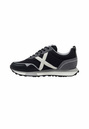 Zapatilla negra y gris con diseño de cruz blanca en el lateral, cordones negros, talón gris y suela texturizada para mayor tracción.