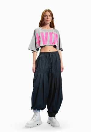 Femme debout portant un t-shirt court gris avec des lettres roses, un pantalon large noir, des bottes blanches épaisses, sur un fond blanc uni.