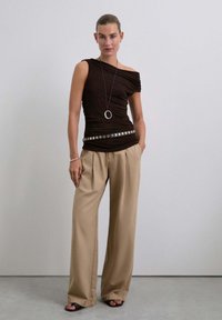 Mujer vestida con una blusa negra de un solo hombro, pantalones beige de pierna ancha, cinturón con tachuelas, un collar largo con colgante y sandalias de tacón negras, de pie contra una pared blanca.