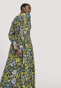Frau mit kurzem Haar trägt ein langärmliges Maxikleid mit grünen, blauen, gelben und pinken Blumenmustern und steht vor einem schlichten hellen Hintergrund.