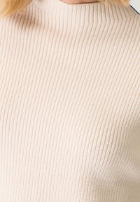 Pull en maille côtelée de couleur beige clair, avec un col montant et une texture douce, mettant en valeur des lignes verticales et une silhouette ajustée.