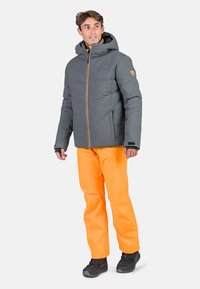 Grau isolierte Jacke mit Kapuze, diagonal genäht und mit goldenen Reißverschluss, kombiniert mit leuchtend orangen Hosen und schwarzen Stiefeln.