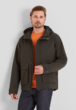 Man wearing dark green hooded jacket over orange sweater and dark jeans, standing against a plain light gray background.