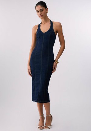 Donna che indossa un vestito midi in denim blu scuro senza maniche con scollo all’americana, orecchini a cerchio dorati, braccialetti e tacchi bianchi con cinturino alla caviglia.