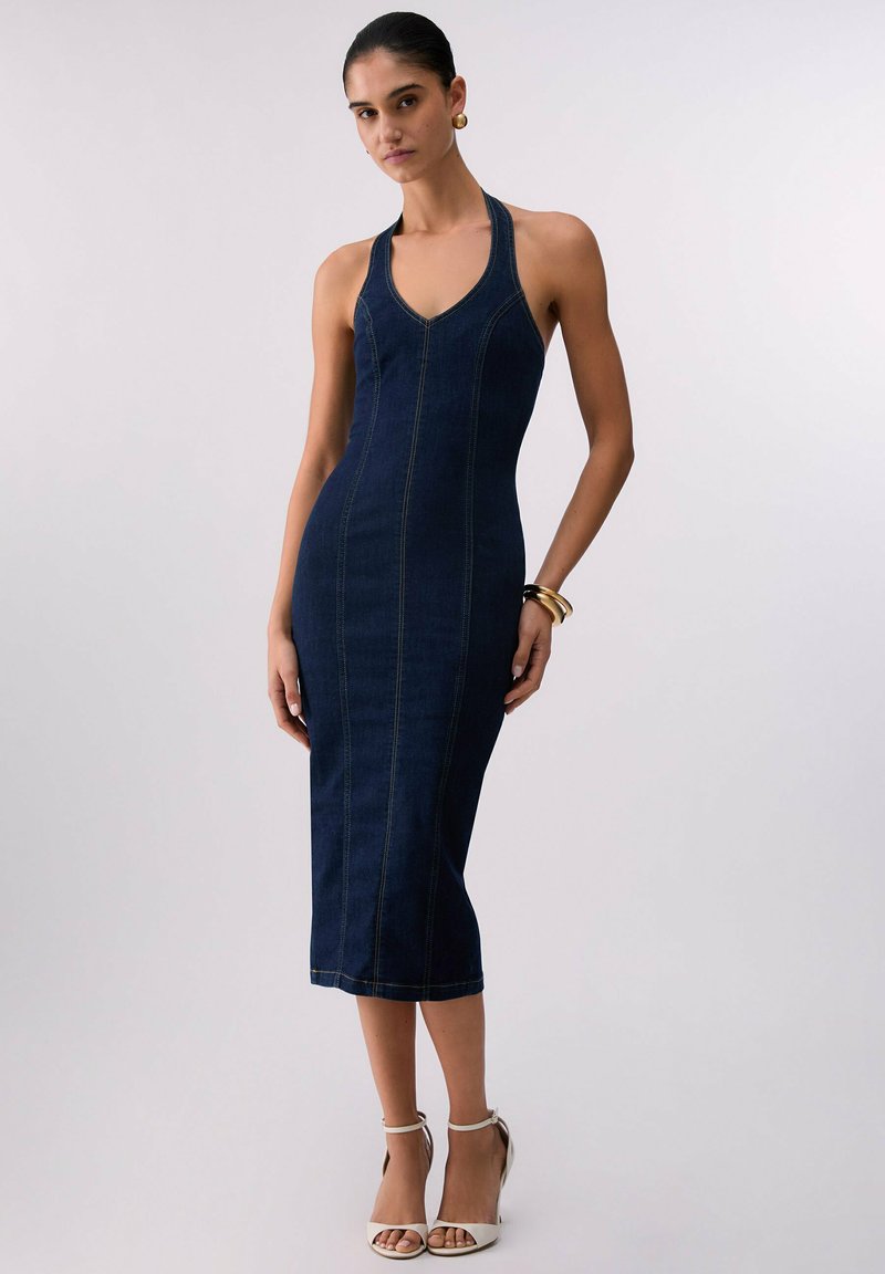 Femme portant une robe midi en denim bleu foncé sans manches avec un col halter, des boucles d'oreilles créoles dorées, des bracelets et des talons blancs à bride à la cheville.