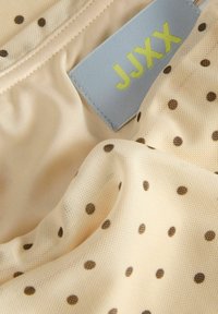 Tissu crème à pois marron avec une étiquette bleue indiquant le nom de la marque "JJXX" en lettres jaunes.