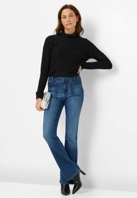 Maglione lungo a maniche lunghe nero e scintillante abbinato a jeans a zampa blu; tiene in mano una clutch argentata; indossa stivali neri con tacco.