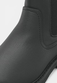 Botă ankle din piele neagră, cu un finisaj texturat, panouri elastice pe laterale și o talpă robustă. Vedere de apropriere care evidențiază detalii și formă.